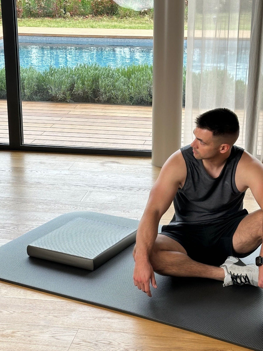 Airex Balance-Pad Elite grau auf Trainingsmatte, Mann sitzt daneben vor Dehnübung, Gleichgewichtstraining