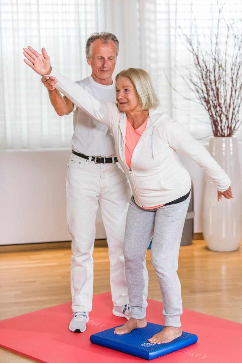 Airex Balance-pad Solid blau, Patientin übt einbeinigen Stand mit Therapeutin-Unterstützung zur Gleichgewichtsschulung